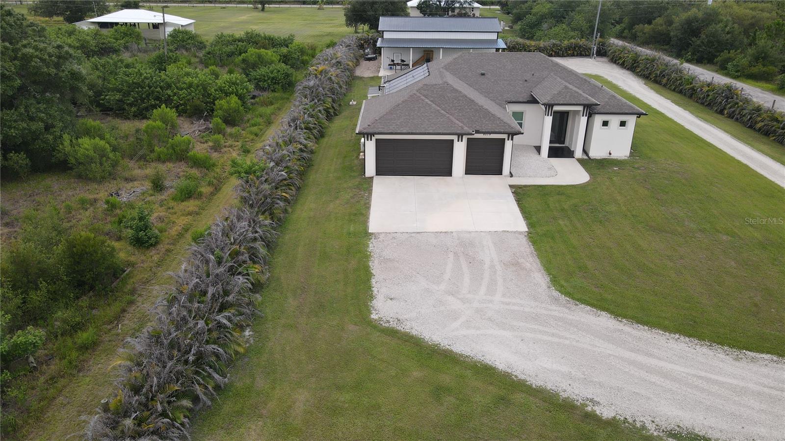 High-altitude drone overview illustrating the true scale and impressive privacy of this custom compound. Lined completely by dense, mature tropical privacy hedges, the estate boasts a grand gravel driveway turnaround, expansive cleared green spaces, and a perfect configuration between the primary residence and the massive detached outbuilding workshop.