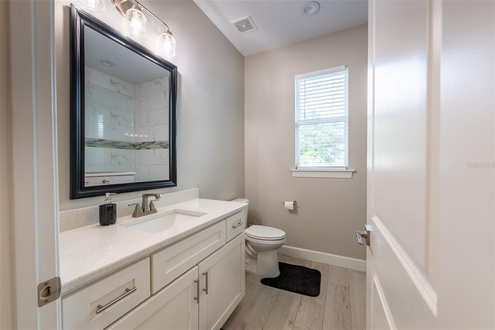 This pristine guest bathroom is loaded with premium finishes, boasting an oversized single vanity with custom white shaker cabinetry, elegant brushed nickel hardware, a sparkling quartz countertop, uniform tile flooring, and a bright window.