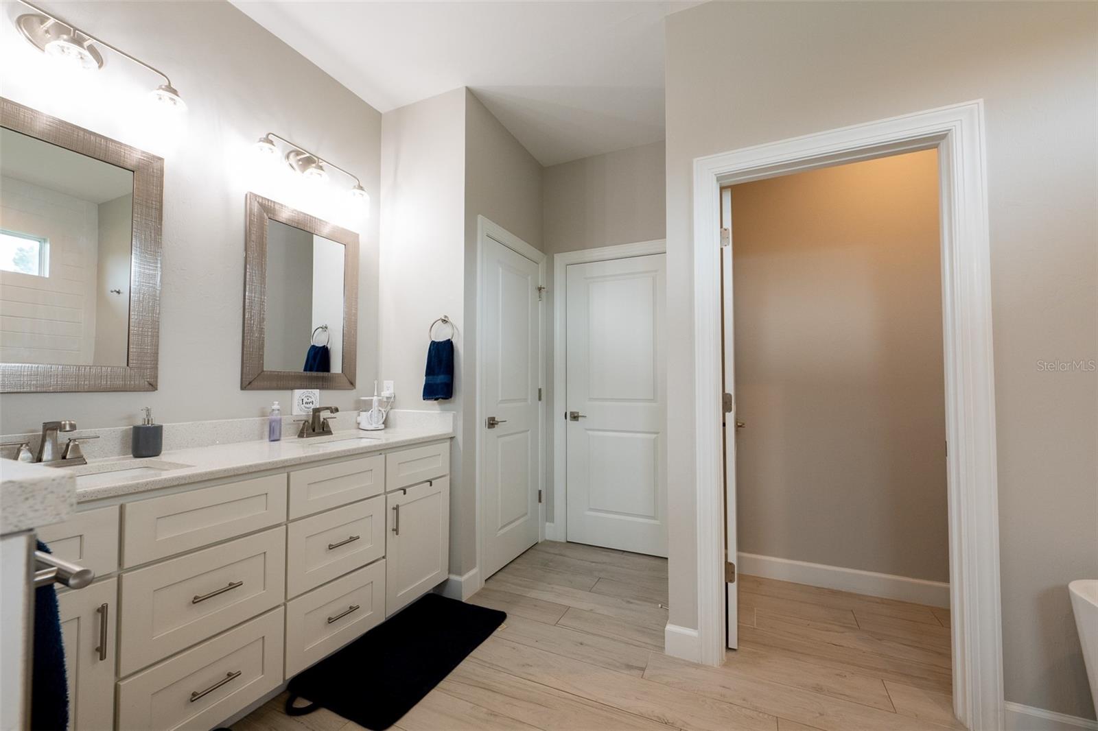 Reverse view of the primary vanity showing the seamless flow of the master suite. High ceilings, uniform wood-look tile flooring, and clean neutral tones lead directly to a private water closet and the expansive walk-in master wardrobe.