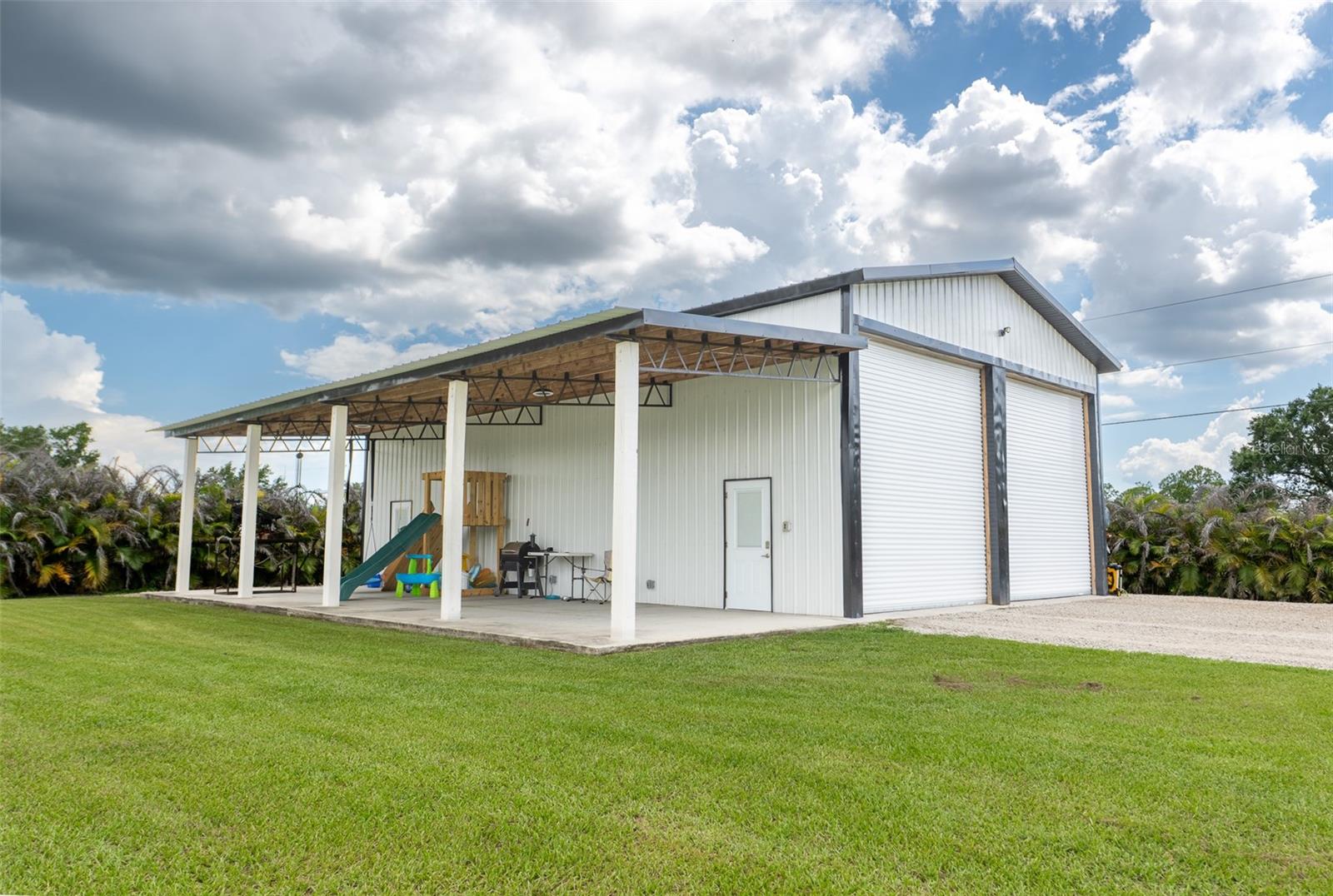 The ultimate hobbyist setup! This massive detached steel workshop/garage features dual high-clearance roll-up doors and a huge covered lean-to/patio area—perfect for an outdoor kitchen, boat storage, or a massive covered play space.