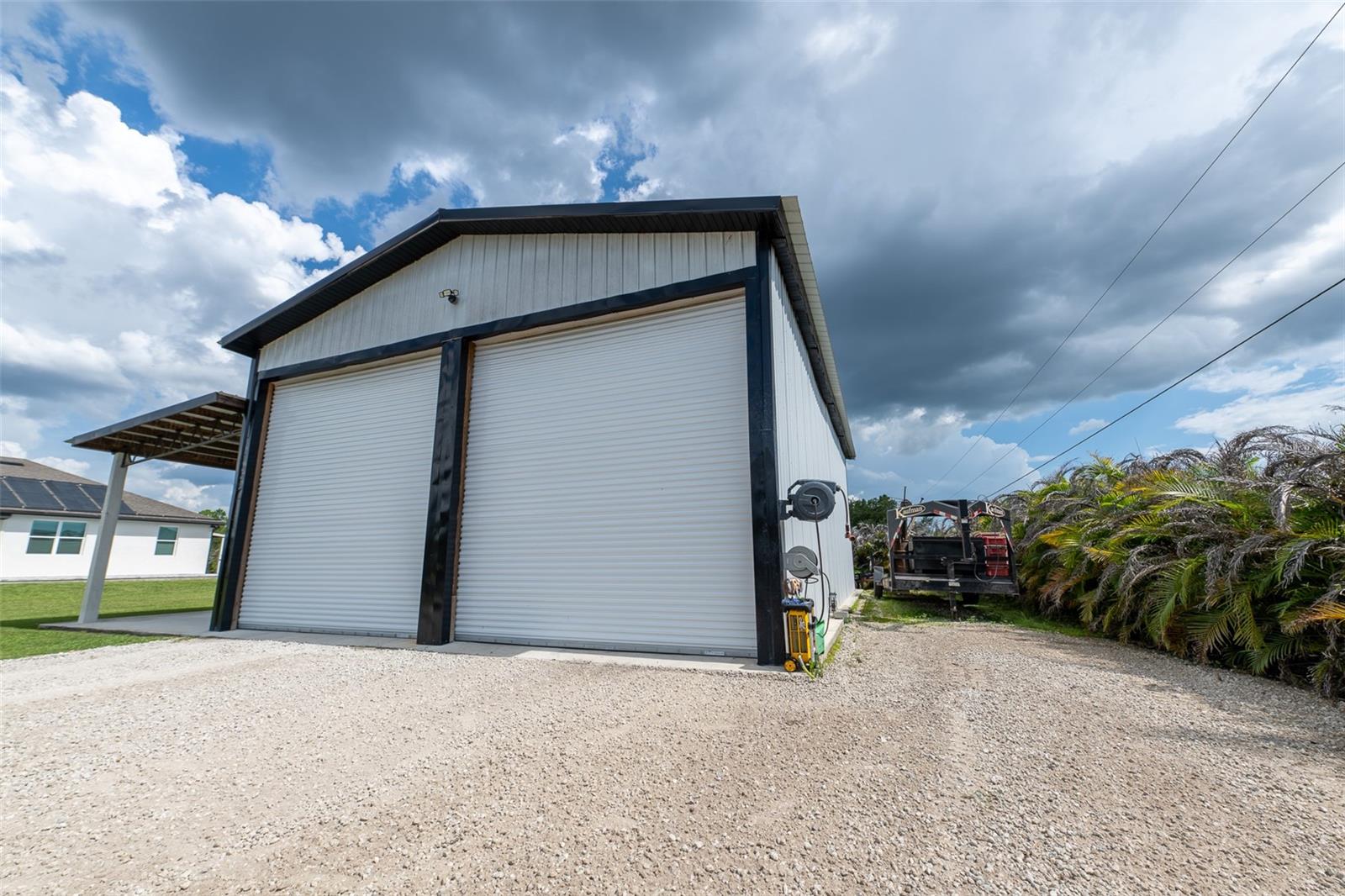 Heavy-duty exterior view of the detached workshop’s commercial-grade, dual roll-up garage doors. Designed for easy drive-in access for oversized trucks, RVs, or boats, with a wide gravel driveway offering plenty of heavy equipment turnaround space.