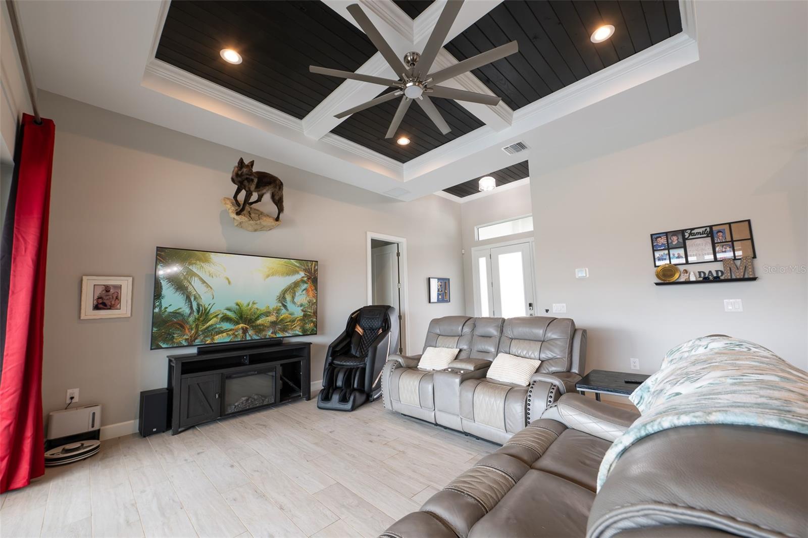 The spacious great room defines modern luxury with its soaring custom tray ceiling, contrasting dark wood shiplap accent, and a massive industrial ceiling fan. The bright, open-concept living area flows effortlessly, making it the perfect central gathering space.