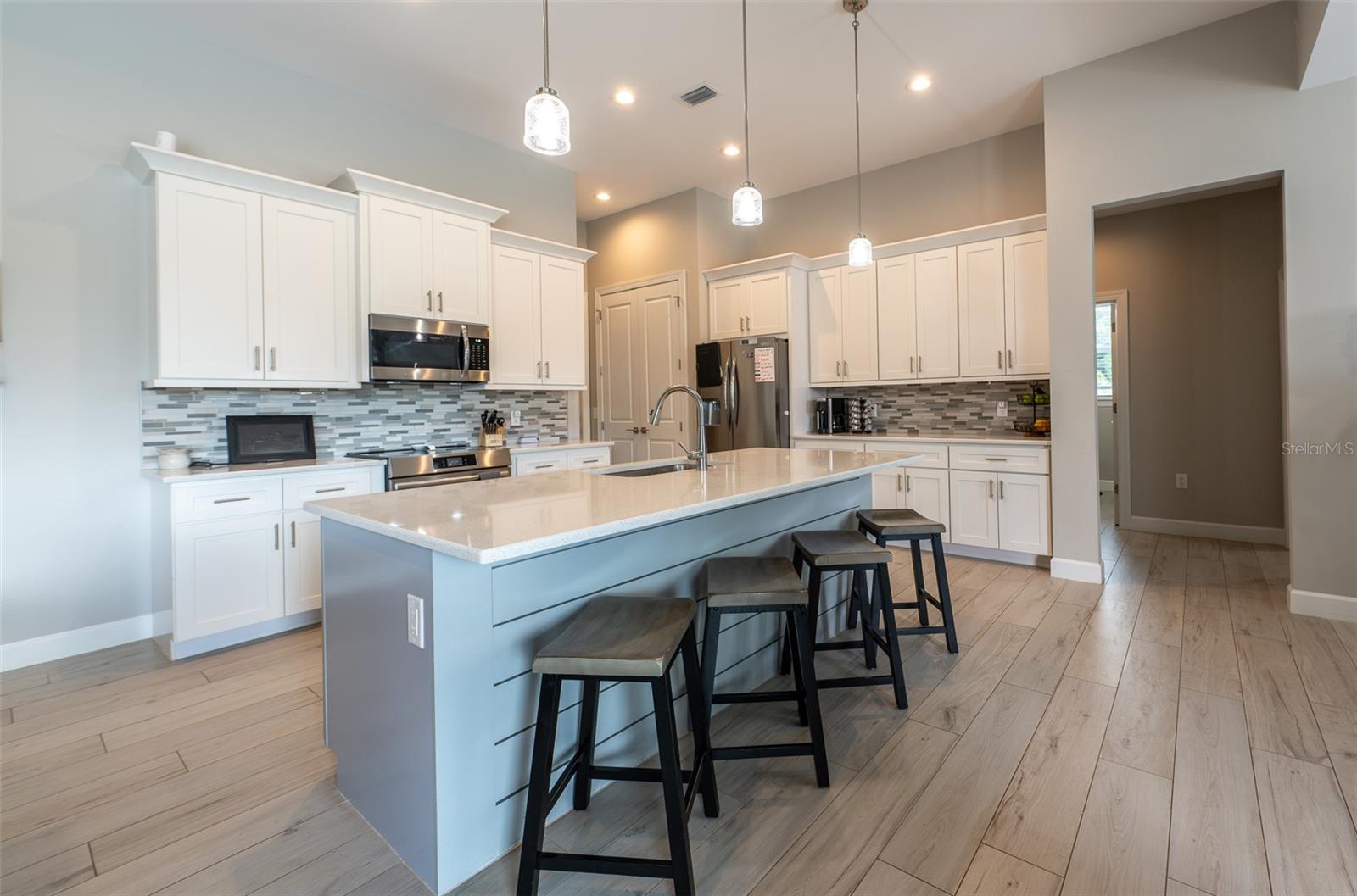 A chef's dream! This gourmet kitchen boasts a massive central island with stylish shiplap detailing and breakfast bar seating, pristine quartz countertops, timeless white shaker cabinets, premium stainless steel appliances, and elegant glass pendant lighting.